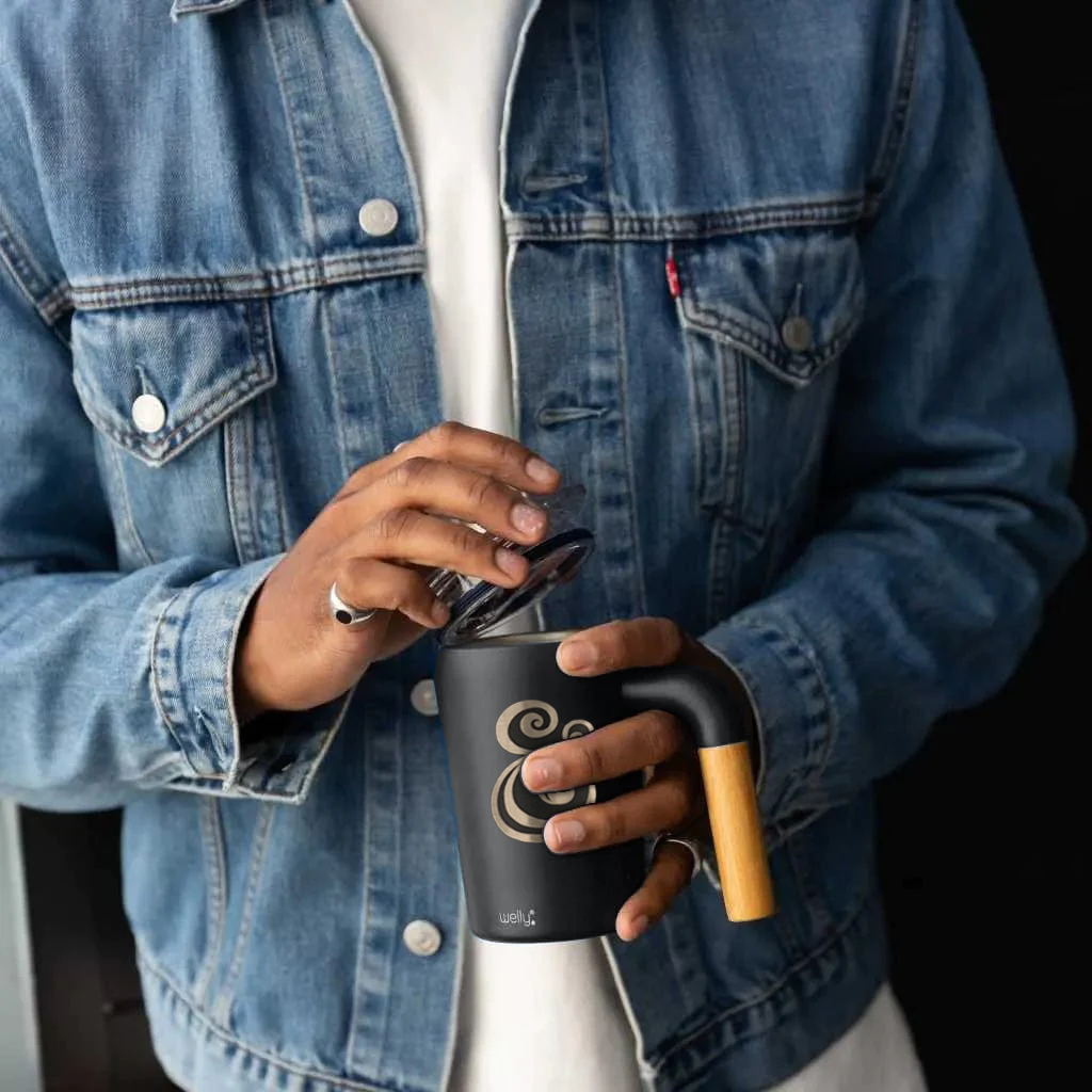 A person wearing a jean jacket holds an insulated mug with the ampersand etched on the side.